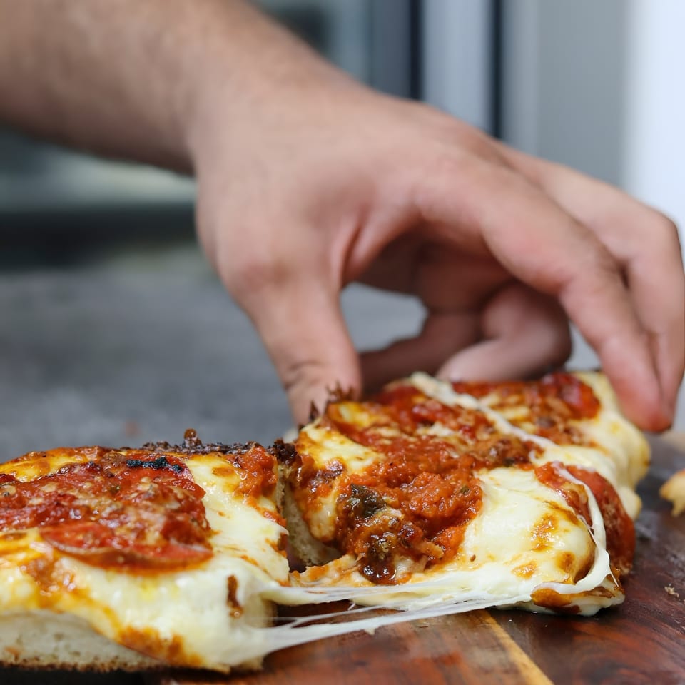A hand pulls a slice of Valentino's pepperoni pizza, stretching the melted cheese.